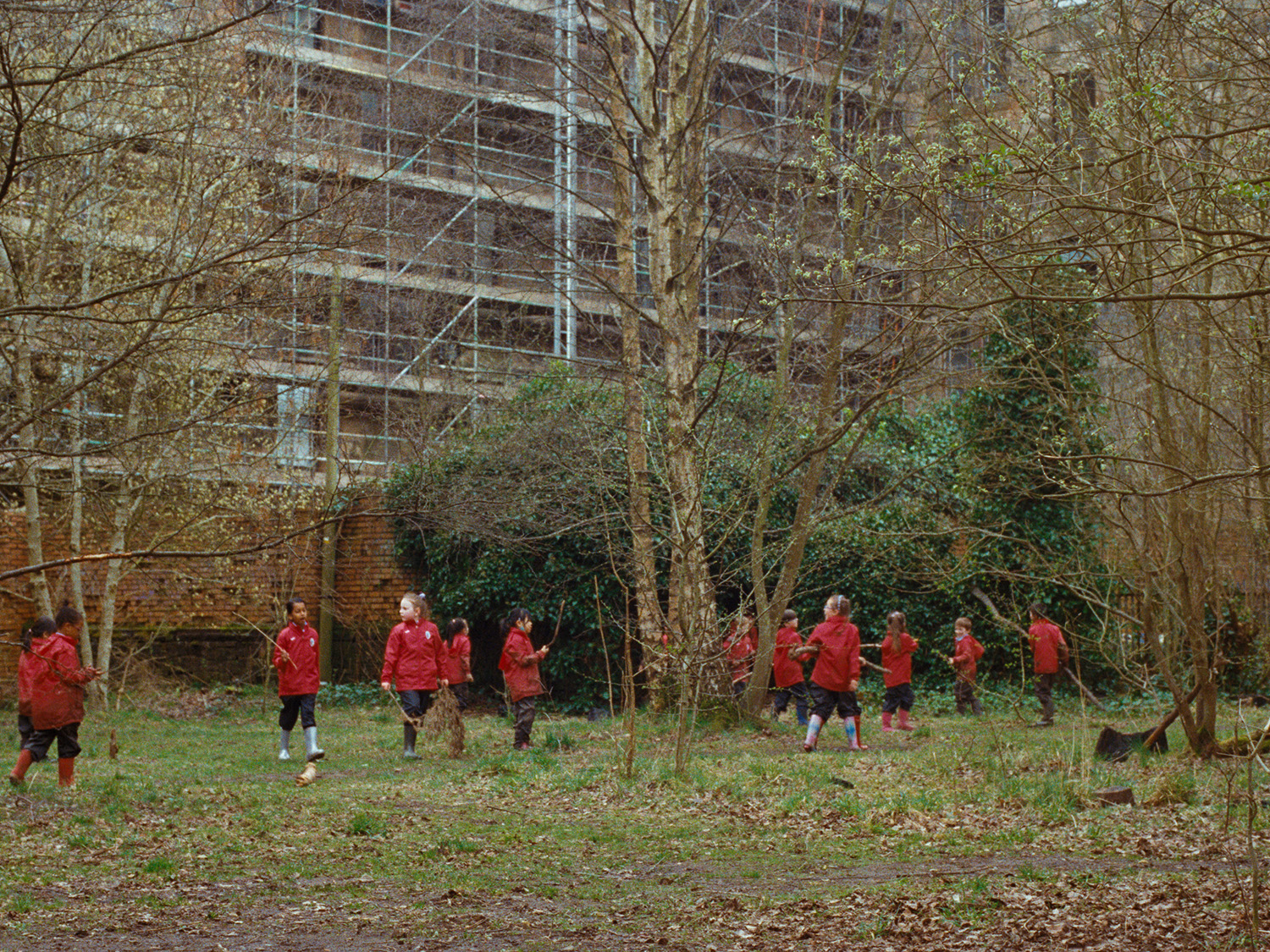We know a better word than happy, video still, children in red, playing in woodland below tall tenement buildings with scafolding